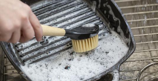 A person uses the scrub brush to wash a soapy Lodge Cast Iron Grill Pan in the sink.